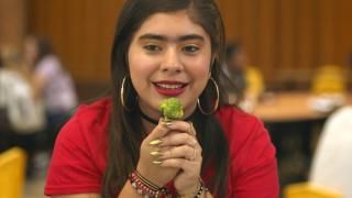 SKAM Austin-serien: En teenagepige iført en rød skjorte, store sølvringe øreringe og flere armbånd sidder i en skolecafeteria med et stykke broccoli mod læberne.