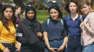 SKAM Austin-serien: Fem teenagepiger står sammen i en linje på en skolegrøn plet og ser trodsigt på kameraet.