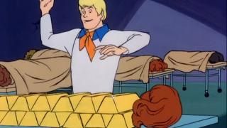 Scooby Doo šovs (TV): Freds stāv blakus morga eksāmenu galdam ar rokām gaisā un pārsteigtu seju; zem palaga ķermeņa vietā bija zelta stieņi.