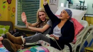 Alexa & Katie seeria: Kaks teismelist tüdrukut lebavad kõrvuti haiglavoodis, naeravad ja tõstavad õhku ilmekalt käsi.