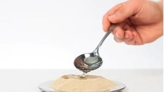 Uma mão segurando uma colher derrama um líquido claro em uma substância arenosa em um prato branco.