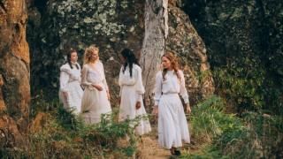 Piknikas „Hanging Rock“ serijoje: keturių jaunų moterų grupė stovi po medžiu šalia didelės uolos; visi dėvi baltas XIX amžiaus sukneles.