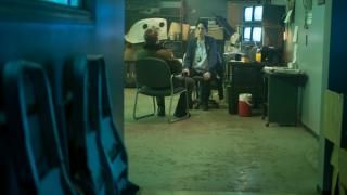 The Crossing TV Series: В слабо осветена стая мъж и жена седят един срещу друг на столове и говорят за нещо, което очевидно е тревожно.